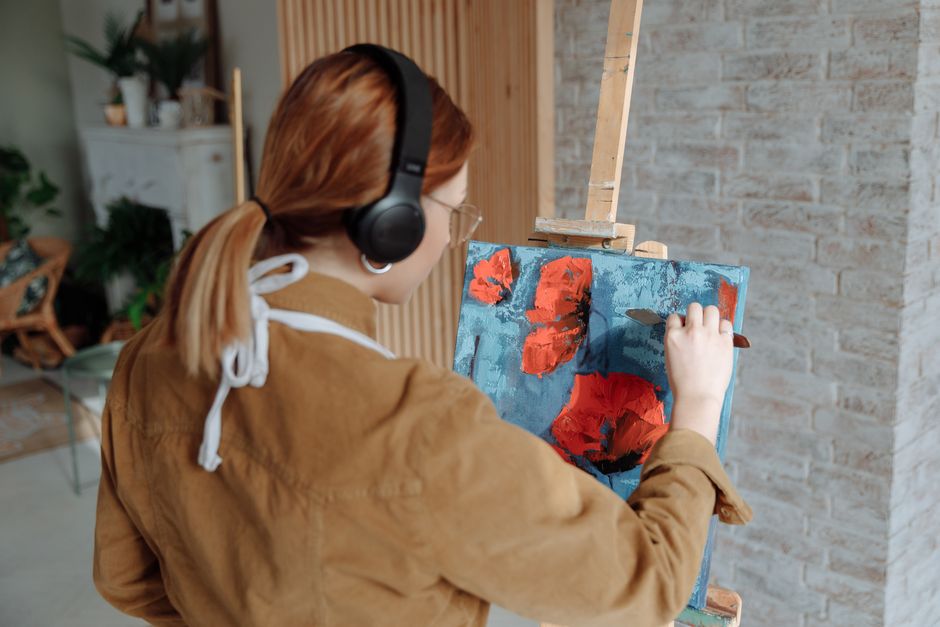 Adult woman using palette knife for painting on canvas with wireless headphones in a cozy studio.