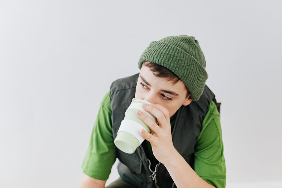 Teenager in a green beanie and shirt drinking from a cup on a white background.